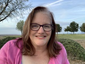 Woman with brown hair and glasses sitting in a park, enjoying the blue sky and trees before she gives an inspiring talk.