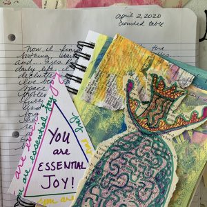 A notebook on a table with an art journal with a variety of small paper works of art. One is a queenly figure, another is a triangle with a square that reads, "You are essential joy" from a poem by Hildegard de Bingen